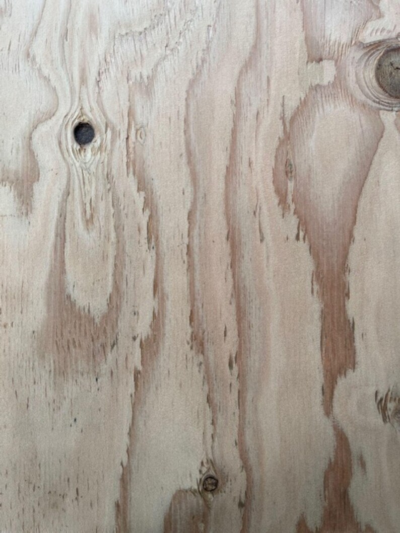 May include: A light-coloured plywood sheet featuring a natural wood grain pattern with visible vertical streaks in shades of tan and beige. The surface displays several dark circular knots of varying sizes scattered across the textured wooden panel.