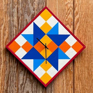 May include: A diamond-shaped wall clock featuring a geometric quilt-inspired pattern in shades of blue, orange, yellow, and white. The clock has a thin red frame and three black hands positioned at the centre of the star-like design.
