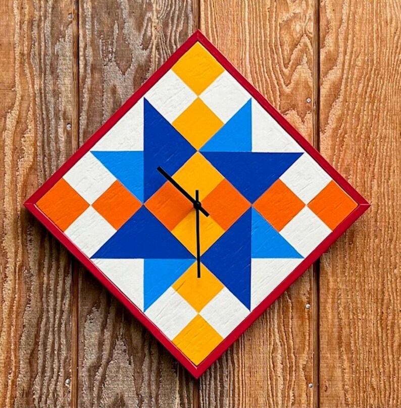 May include: A diamond-shaped wall clock featuring a geometric quilt-inspired pattern in shades of blue, orange, yellow, and white. The clock has a thin red frame and three black hands positioned at the centre of the star-like design.