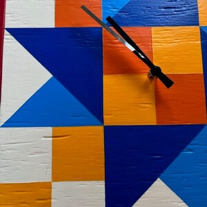 May include: A square wall clock featuring a geometric quilt-inspired pattern painted in vibrant shades of orange, white, royal blue, and light blue. The clock face has a textured wood grain surface and two thin black hands centred on the design.