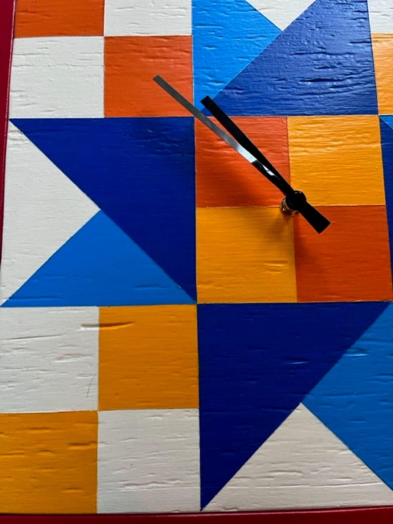 May include: A square wall clock featuring a geometric quilt-inspired pattern painted in vibrant shades of orange, white, royal blue, and light blue. The clock face has a textured wood grain surface and two thin black hands centred on the design.