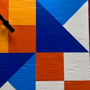 May include: A square wall clock featuring a geometric pattern of painted wooden blocks in vibrant shades of orange, yellow, white, and blue. The clock face displays a grid of triangles and squares with black clock hands extending from the centre.