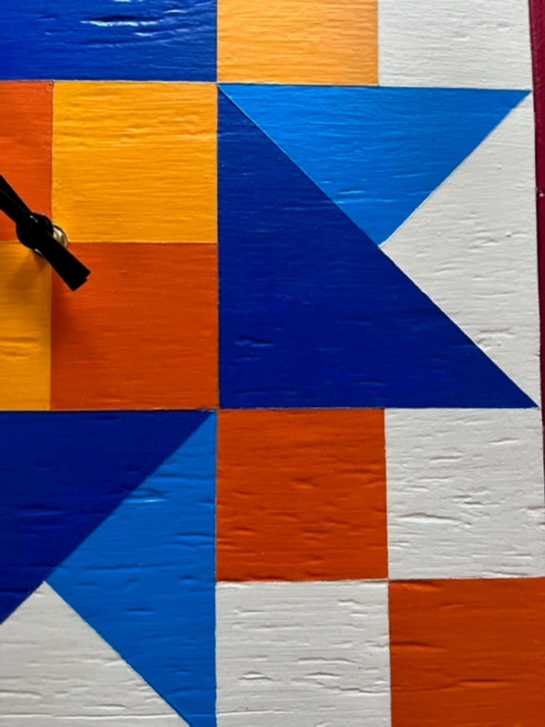 May include: A square wall clock featuring a geometric pattern of painted wooden blocks in vibrant shades of orange, yellow, white, and blue. The clock face displays a grid of triangles and squares with black clock hands extending from the centre.