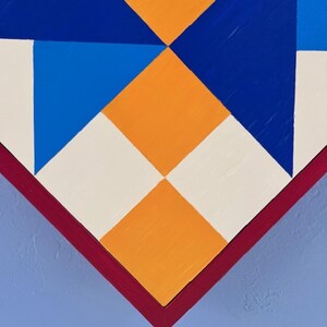 May include: A geometric wall art piece featuring a pattern of triangles and squares in vibrant orange, deep blue, bright blue, and off-white. The composition is framed by a dark red border and set against a solid light blue background surface.