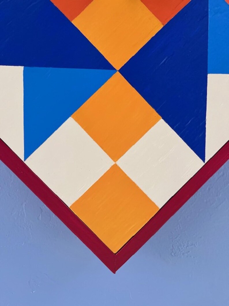 May include: A geometric wall art piece featuring a pattern of triangles and squares in vibrant orange, deep blue, bright blue, and off-white. The composition is framed by a dark red border and set against a solid light blue background surface.