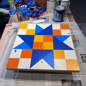 May include: A square wooden board featuring a painted geometric star pattern in shades of bright blue, orange, and white. The design consists of a central four-square orange block surrounded by blue triangular points and alternating orange and white squares.