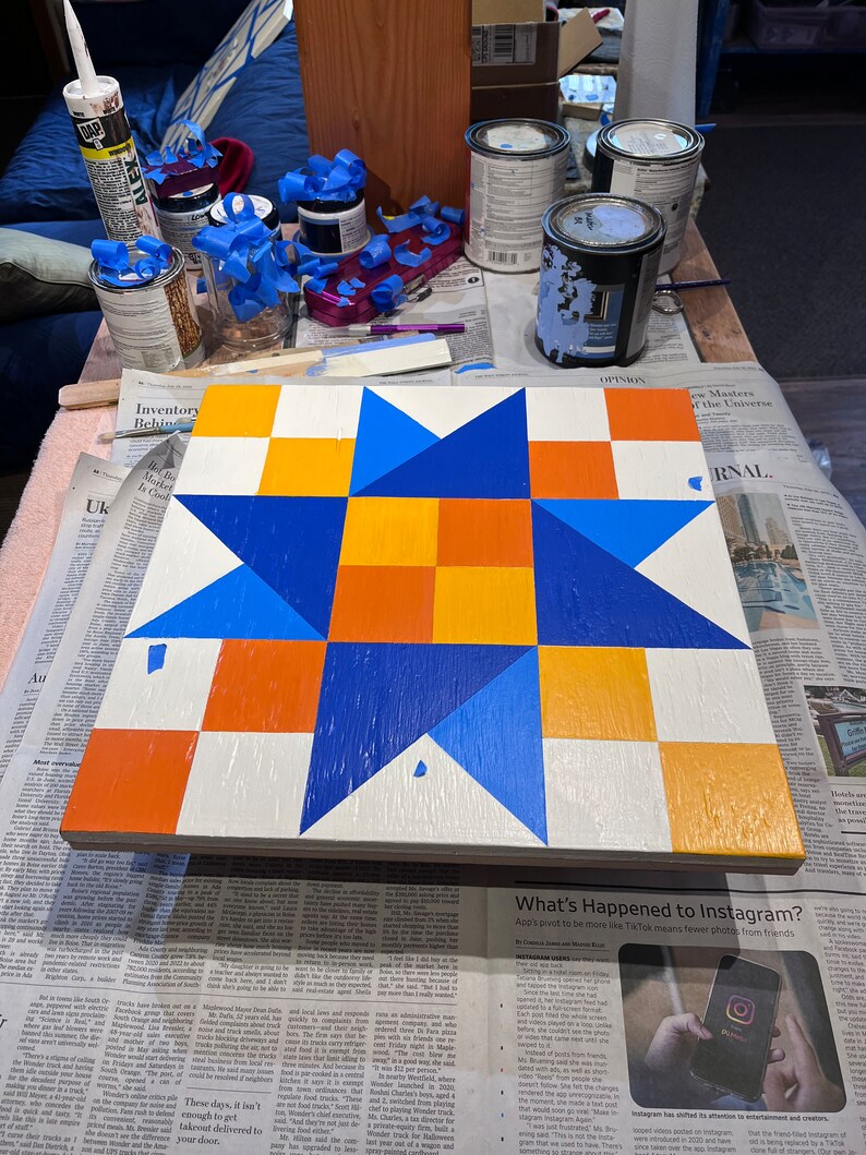 May include: A square wooden board featuring a painted geometric star pattern in shades of bright blue, orange, and white. The design consists of a central four-square orange block surrounded by blue triangular points and alternating orange and white squares.