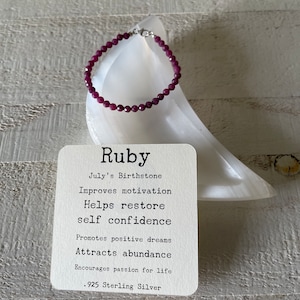 May include: A red ruby gemstone bracelet with a sterling silver clasp. The bracelet is on a white surface with a card that reads "Ruby, July's Birthstone, Improves motivation, Helps restore self confidence, Promotes positive dreams, Attracts abundance, Encourages passion for life, .925 Sterling Silver."