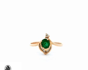 May include: A gold ring featuring a vibrant green emerald gemstone at the center, surrounded by smaller, sparkling diamonds. The ring has a unique, leaf-like design with delicate details and a polished finish. The ring is set against a white background.