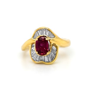 Puede incluir: Un anillo de oro con una piedra preciosa de rubí rojo rodeada por un halo de diamantes talla baguette.