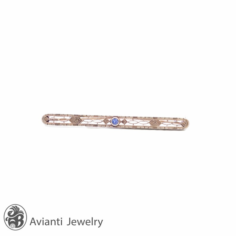May include: A delicate gold bar pin with a blue gemstone in the center. The pin is made of intricate filigree work and has a geometric design.