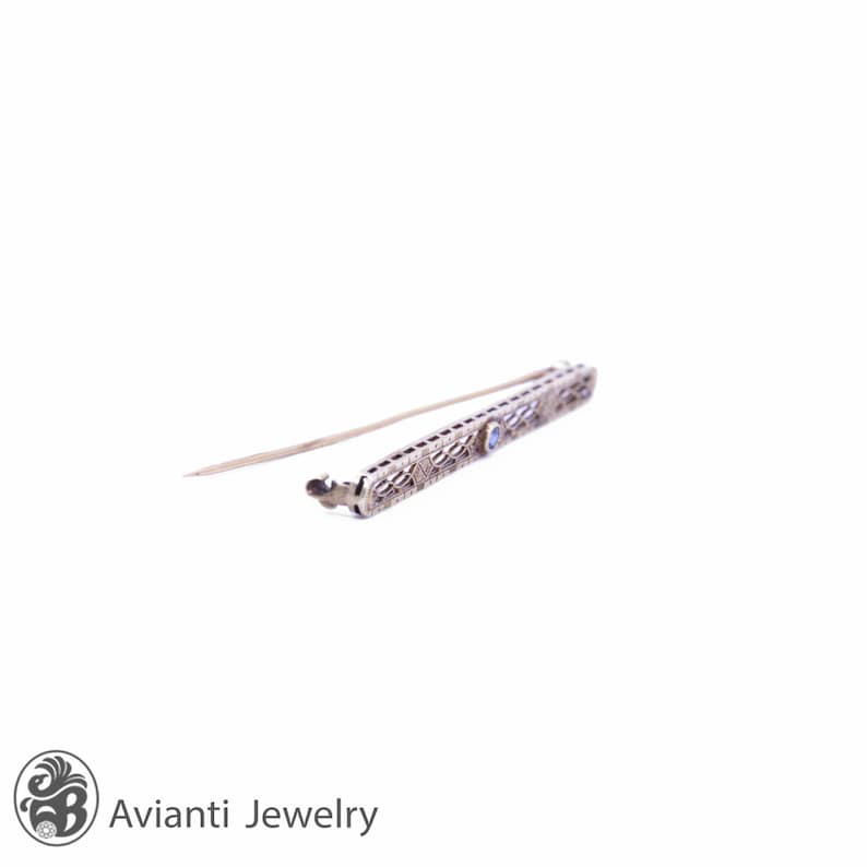 May include: A delicate silver filigree bar brooch with a single blue gemstone in the center.
