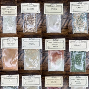May include: A collection of 12 small plastic bags filled with different spice blends. Each bag has a label with the name of the blend, such as "Bacon", "Onion", "Garlic", "Sun-Dried Tomato", "Garlic Jalapeno", "Dill", "Jalapeno", "Chipotle Ranch", "Spinach", "Bacon Ranch", "Buffalo Ranch", "Horseradish Bacon", "Fiesta", and "Creamy Cucumber".