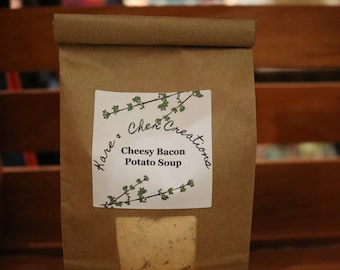 Op de afbeelding: Een bruine papieren zak met een wit label waarop "Hare & Cheri Creations" en "Cheesy Bacon Potato Soup" staat. De zak ligt op een houten oppervlak.