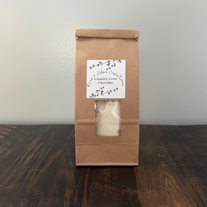 May include: A brown paper bag with a white label that says "Here's Cher Creations Country Corn Chowder". The bag has a clear window showing the contents of the bag.
