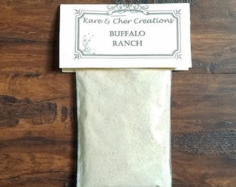 Op de afbeelding: Een klein, verzegeld zakje Buffalo Ranch kruidenmix ligt op een houten oppervlak. Een wit etiket boven het zakje leest "Kare & Cher Creations" en "BUFFALO RANCH". Het zakje bevat een lichtgekleurd poeder.
