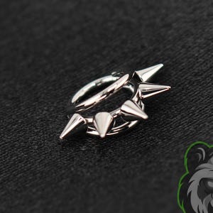 May include: A silver-toned circular hoop earring featuring four evenly spaced, pointed spikes along the outer edge. The metal has a polished, reflective finish. A logo depicting a stylized panda face with the text PIERCING PANDA is visible.