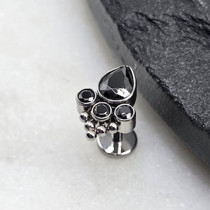 May include: A silver-toned, decorative piercing stud featuring a cluster of black gemstones. The design includes a large, teardrop-shaped stone and several smaller, round stones. The stud is set against a white and black background.