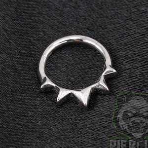 May include: A silver-colored circular hoop ring featuring a series of five pointed triangular spikes along the bottom half of the band. The metal has a polished, reflective finish, and the design creates a sunburst or crown-like appearance.