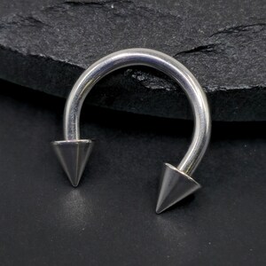 May include: A silver horseshoe piercing with pointed ends. The curved bar has two conical tips. The jewellery is made of a shiny metal and is set against a dark, textured background.