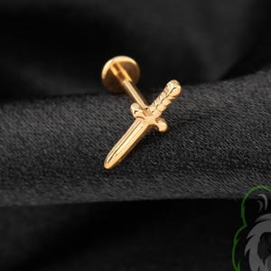 May include: A gold-toned metal stud earring shaped like a small sword with a textured hilt and a pointed blade. The jewelry piece features a flat circular backing post attached to the center of the sword design, presented in a polished finish.