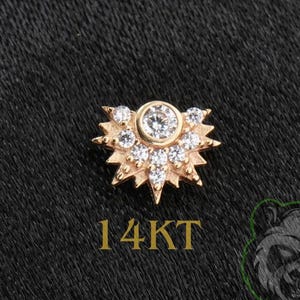 May include: A gold body jewelry piece featuring a fan-shaped design with a central round clear stone surrounded by smaller clear stones in a sunburst pattern. The text 14KT is printed in gold below the jewelry piece on a black textured background.
