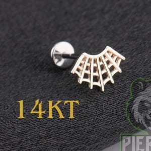 May include: A 14KT gold labret piercing stud featuring a decorative top shaped like a spider web. The jewelry includes a flat circular back disc and a post, with the text 14KT visible in yellow lettering near the bottom left of the image.