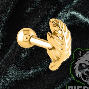 May include: A gold-colored ear piercing stud with a leaf design. The stud features a small ball on one end and a detailed leaf on the other. The background is a dark green, textured fabric.