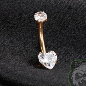 May include: A curved barbell body jewelry piece featuring a rose gold-toned metal shaft, a small round clear gemstone at the top, and a larger heart-shaped clear gemstone at the bottom. The jewelry is displayed against a dark fabric background.