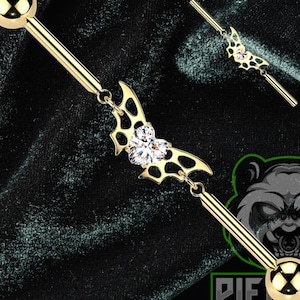 May include: Gold-colored industrial barbell with a bat-shaped charm and a clear gem. The barbell has a ball on each end and a decorative charm with a clear gem in the center. The background is a dark green velvet.