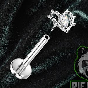 Puede incluir: Un piercing de labio plateado con un diseño en forma de corazón. El corazón presenta una gema transparente central y está rodeado por un borde puntiagudo. El perno está separado del poste, que tiene una base de disco plano. El logotipo "Piercing Panda" es visible.