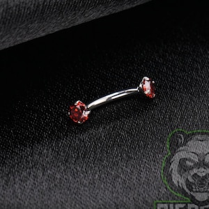 May include: A curved barbell body jewelry piece featuring a polished silver-tone metal shaft with a round, faceted red gemstone set at each end. The jewelry is positioned diagonally, showcasing the vibrant red stones against a dark background.