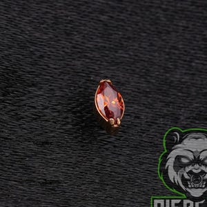 May include: A marquise-cut orange gemstone set in a gold-toned metal prong setting. The stone features a faceted surface that reflects light, creating a vibrant appearance. The piece is designed for body jewelry applications with a small metal base.