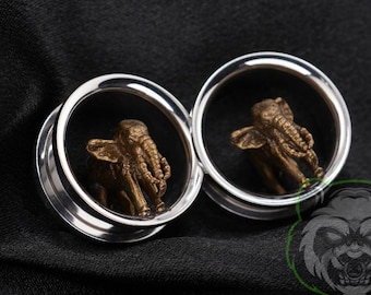 May include: A pair of silver-toned circular ear plugs, each featuring a small, three-dimensional bronze-colored elephant figurine centered within the hollow opening. The metallic tunnels have a smooth, polished finish and a wide rim.