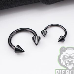 May include: Two black circular barbell piercings with pointed cone ends. The jewellery features a smooth, dark metallic finish and a curved bar design. Each piece is finished with sharp, conical spikes at both ends of the circular hoop structure.
