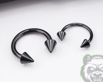 May include: Two black circular barbell piercings with pointed cone ends. The jewelry features a smooth, dark metallic finish and a curved bar design. Each piece is finished with sharp, conical spikes at both ends of the circular hoop structure.