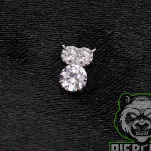 May include: A silver-toned body jewelry piece featuring a small owl-shaped design. The owl's head is formed by two small clear round stones, while its body consists of a single larger clear round stone. The piece has a metallic finish.