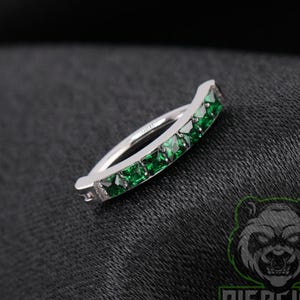 May include: A silver-toned hinged segment ring featuring a row of five square-cut emerald green gemstones set along the outer curve. The jewelry piece has a polished metallic finish and a secure closure mechanism visible on the side.