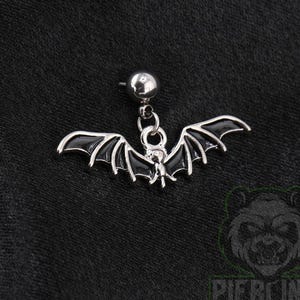 May include: A silver-toned metal stud earring featuring a dangling bat charm. The bat has outstretched wings filled with black enamel detailing and is attached to a small circular stud post with a polished silver finish, creating a cohesive look.