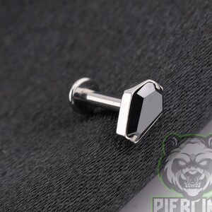 May include: A silver-toned labret piercing stud featuring a flat, coffin-shaped top with a smooth, reflective surface. The jewelry includes a straight post and a circular flat back disc, designed for body modification and cartilage or lip piercings.