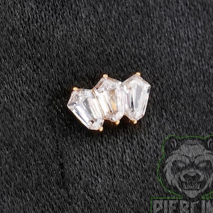 May include: A gold-toned body jewelry piece featuring three clear, faceted, trapezoidal-shaped stones arranged in a cluster. The stones are set in a metallic frame, creating a geometric design with a bright, reflective finish on the clear crystals.