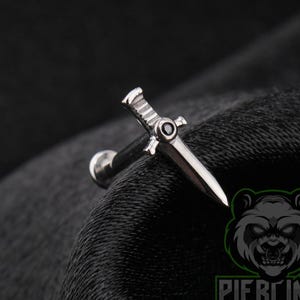 May include: A silver-toned metal body jewelry piece shaped like a small sword or dagger. The hilt features a single small black circular stone set in the center, with a flat, pointed blade extending outward from a straight post and a circular back disc.