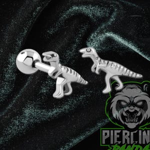 May include: Silver-colored dinosaur-shaped ear piercing jewelry. One piece has a ball on the end, and the other is a standalone dinosaur. The logo "Piercing Panda" is in the lower right corner.
