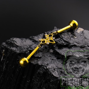 May include: A gold-colored industrial barbell piercing. The barbell features a decorative charm with a star-like design, embellished with small, clear gemstones. The ends of the barbell are rounded. The jewelry is displayed against a dark, textured background.