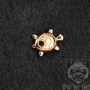 May include: A rose gold-toned metal stud earring shaped like a turtle. The turtle features a detailed, segmented shell pattern on its back, with four legs and a head extending from the central body. The surface has a polished, reflective finish.