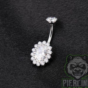 May include: A silver-toned curved barbell belly button ring featuring a large central oval-cut clear gemstone surrounded by a halo of smaller round clear stones, topped with a single small clear round gemstone at the top of the curved post.