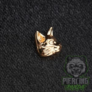 May include: A small, gold-toned metal stud earring shaped like the head of a fox or wolf, featuring pointed ears and detailed facial contours. The piece has a polished, reflective surface and is designed with a three-dimensional profile.