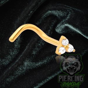 May include: A gold-colored nose stud with a curved post and a three-stone cluster setting. The cluster features three clear, round-cut stones arranged in a triangular pattern at the end of the post, providing a bright, reflective appearance.