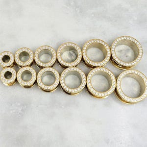 May include: A collection of gold-colored ear gauges with a sparkling, clear crystal border. The gauges are arranged in two rows, showcasing a variety of sizes. The jewelry has a polished finish and a modern aesthetic.
