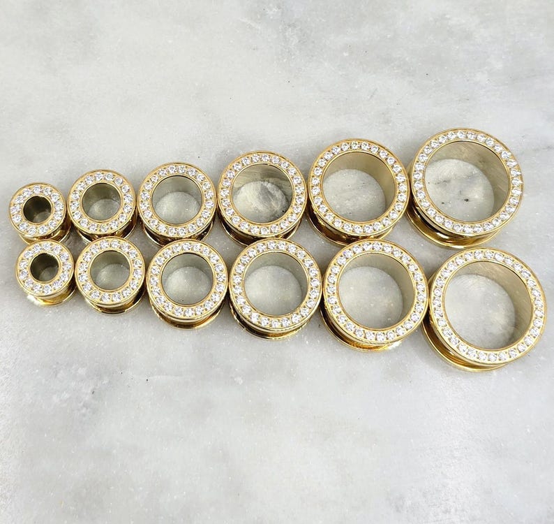 May include: A collection of gold-colored ear gauges with a sparkling, clear crystal border. The gauges are arranged in two rows, showcasing a variety of sizes. The jewelry has a polished finish and a modern aesthetic.
